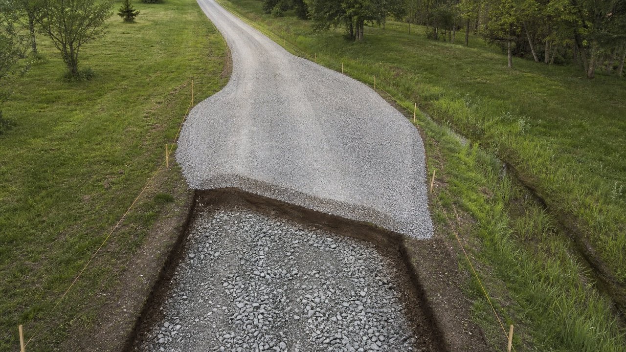 Narrow rural gravel driveway widened into a passing bay with tapered ends, survey stakes, exposed aggregate base, finished gravel, and a drainage swale.