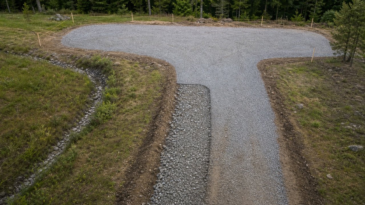 Gravel driveway leading into a broad hammerhead turnaround, with survey stakes, a visible coarse aggregate layer, finished gravel, and a drainage swale.