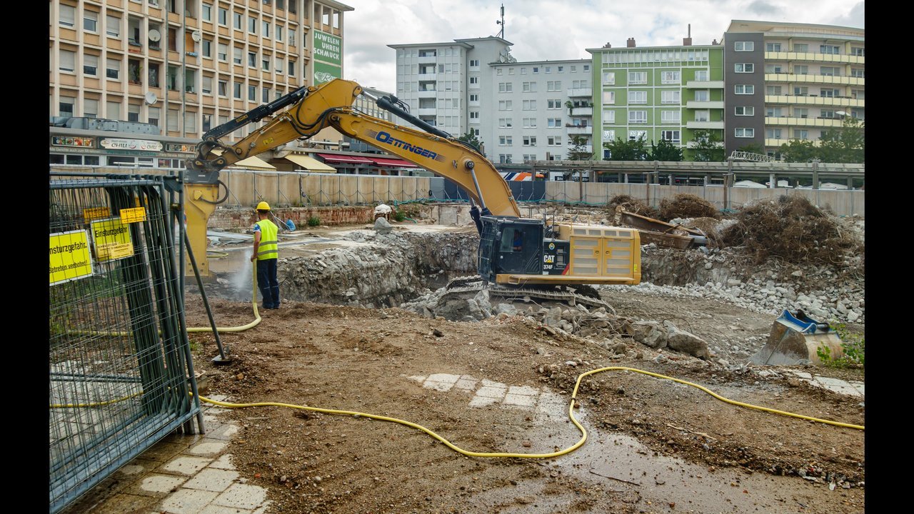 An excavator working inside an open building foundation excavation pit, with a site worker nearby for scale. Photo: Friedrich Haag, CC BY-SA 4.0, via Wikimedia Commons.